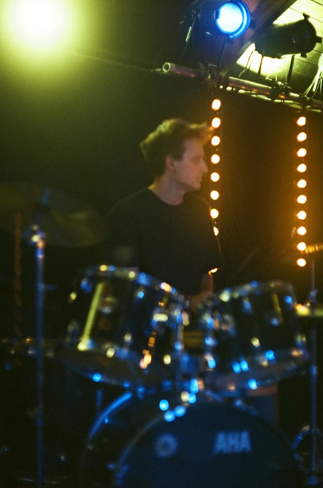 The drummer of Hannetones behind the kit, seen through a cymbal stand under a yellow spotlight